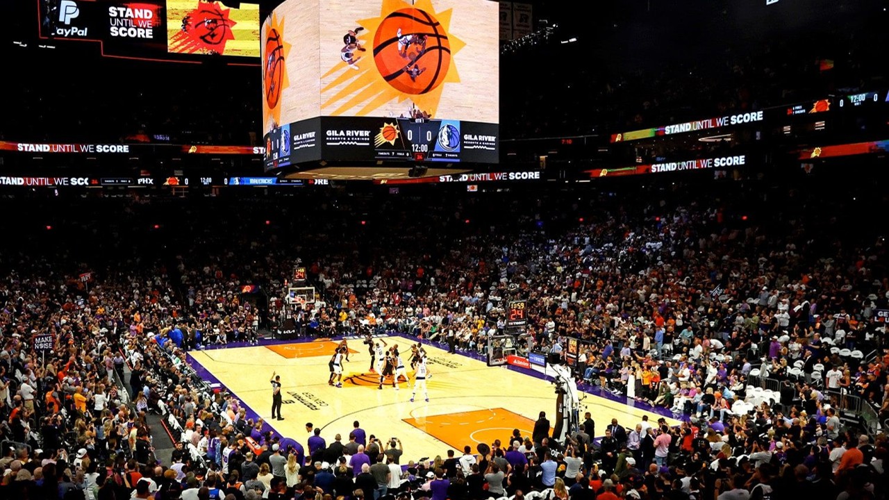 Crowded indoor basketball arena during a game start with players jumping for the opening tip-off under a large center-hung scoreboard showing team logos and scores.