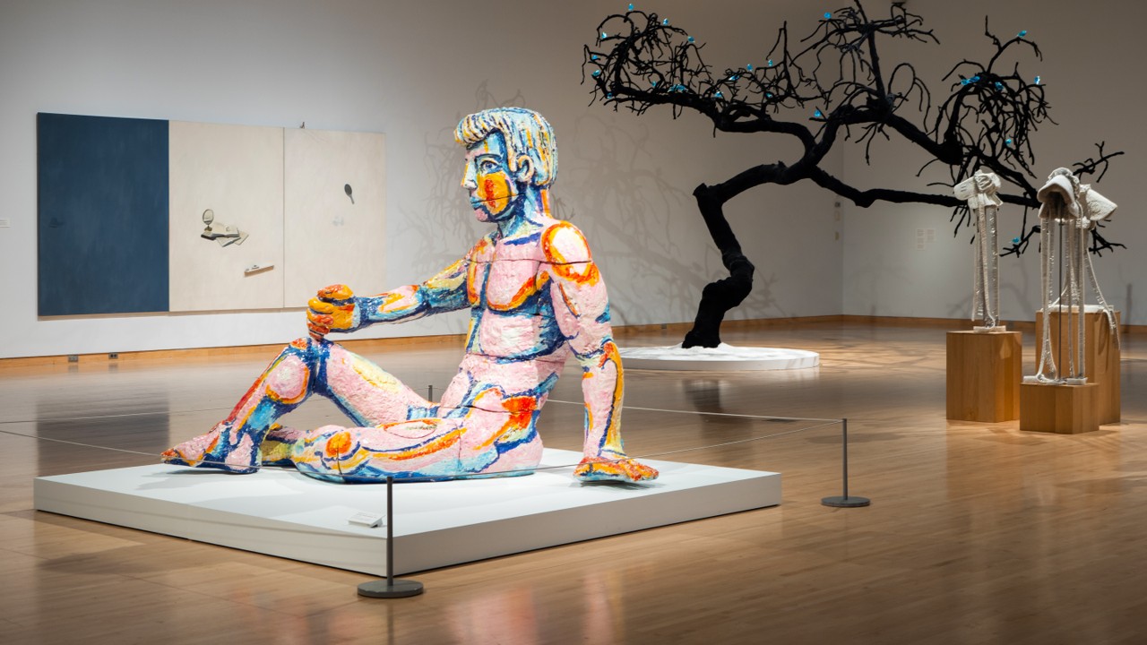Colorful sculpture of a seated human figure with exaggerated muscular lines in blue, pink, orange, and yellow in an art gallery, with a black twisted tree sculpture adorned with blue elements, and two abstract humanoid sculptures on wooden pedestals in the background.