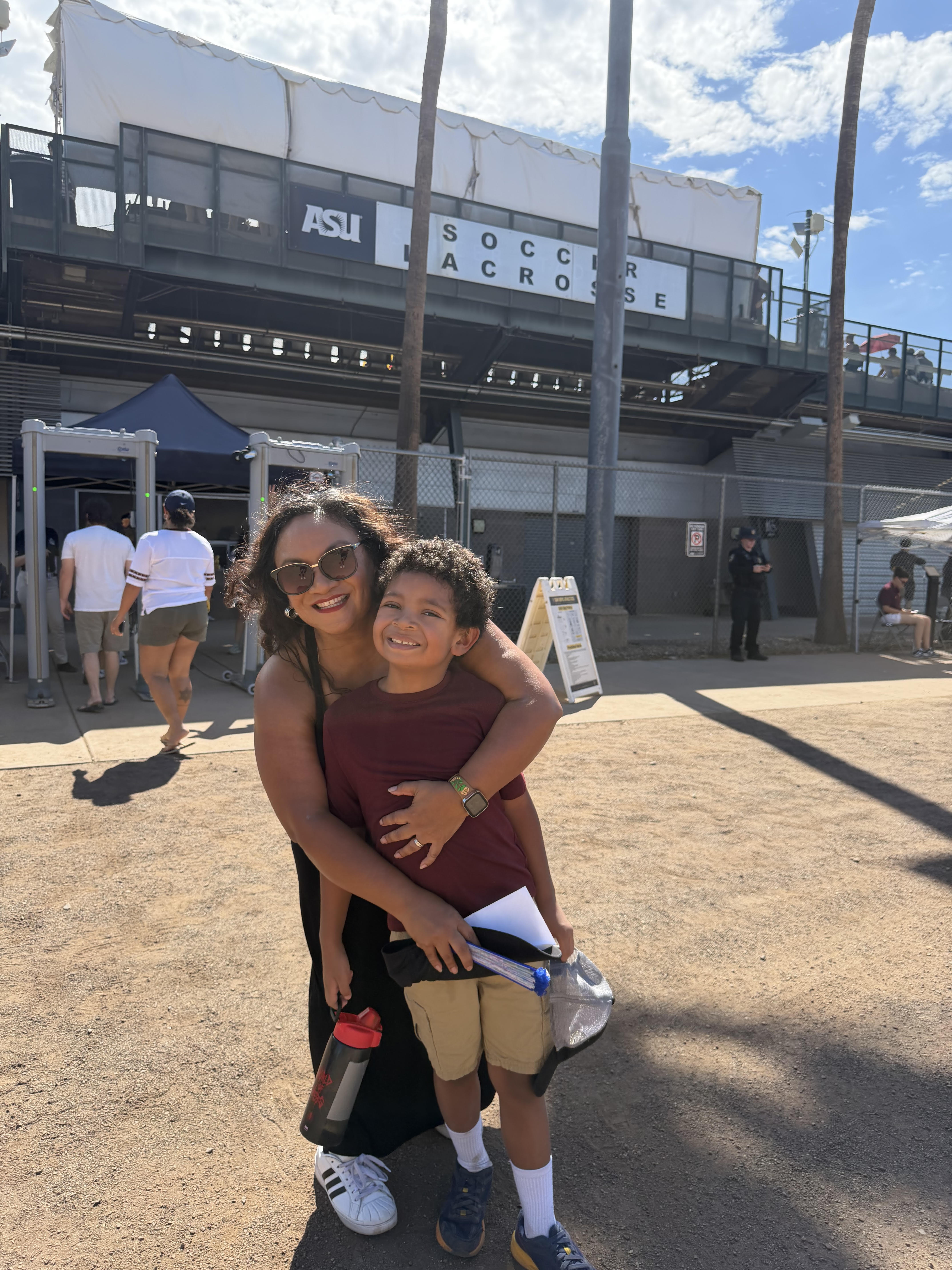 A smiling woman wearing sunglasses and a black dress hugs a smiling young boy in a maroon shirt and beige shorts at an outdoor sports venue with an ASU Soccer Lacrosse sign overhead and a clear blue sky.
