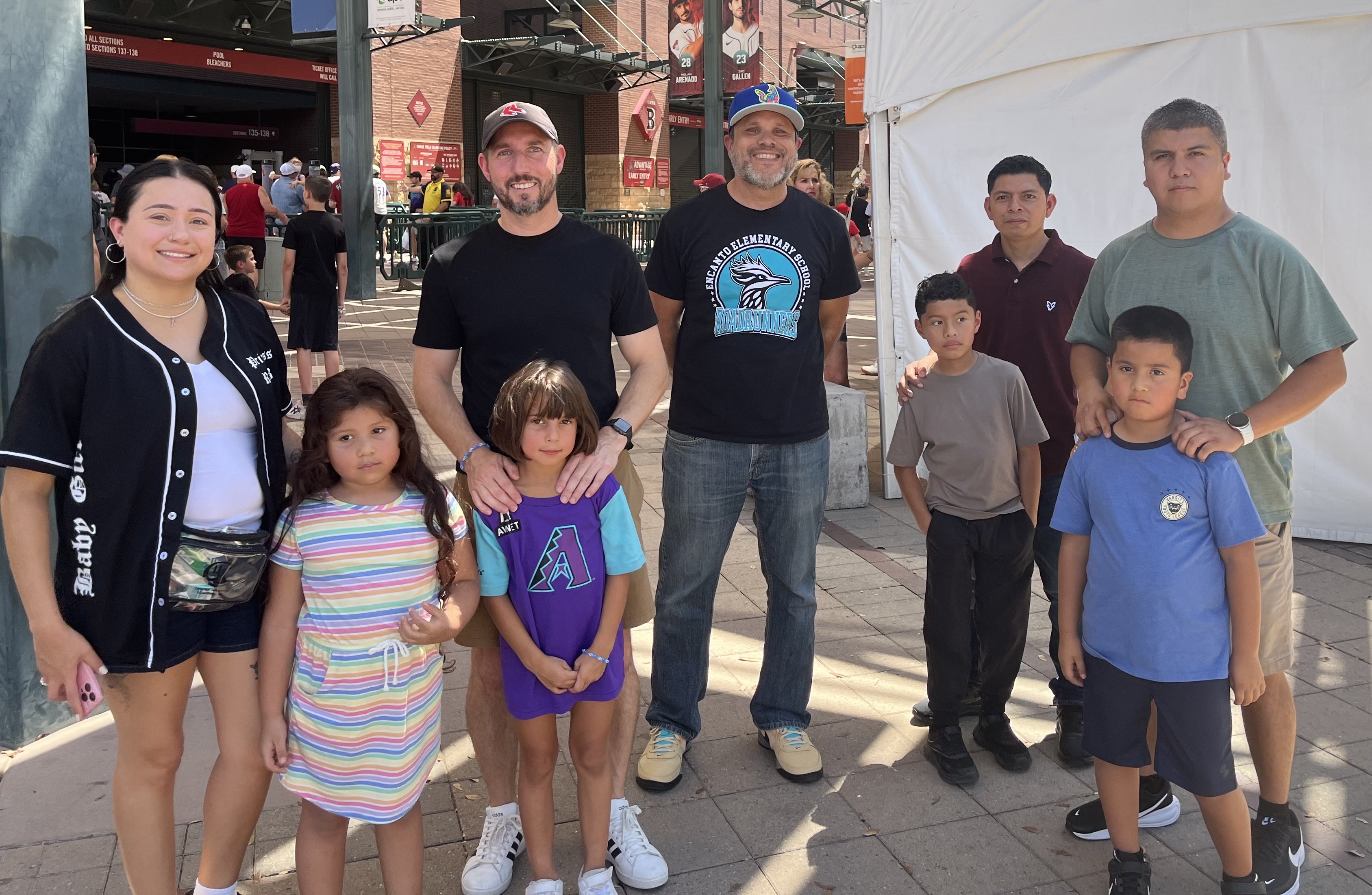 A group of nine people, including four men, one woman, and four children, standing outdoors on a paved area near a stadium entrance, with the men and children dressed casually and the woman holding a pink phone.