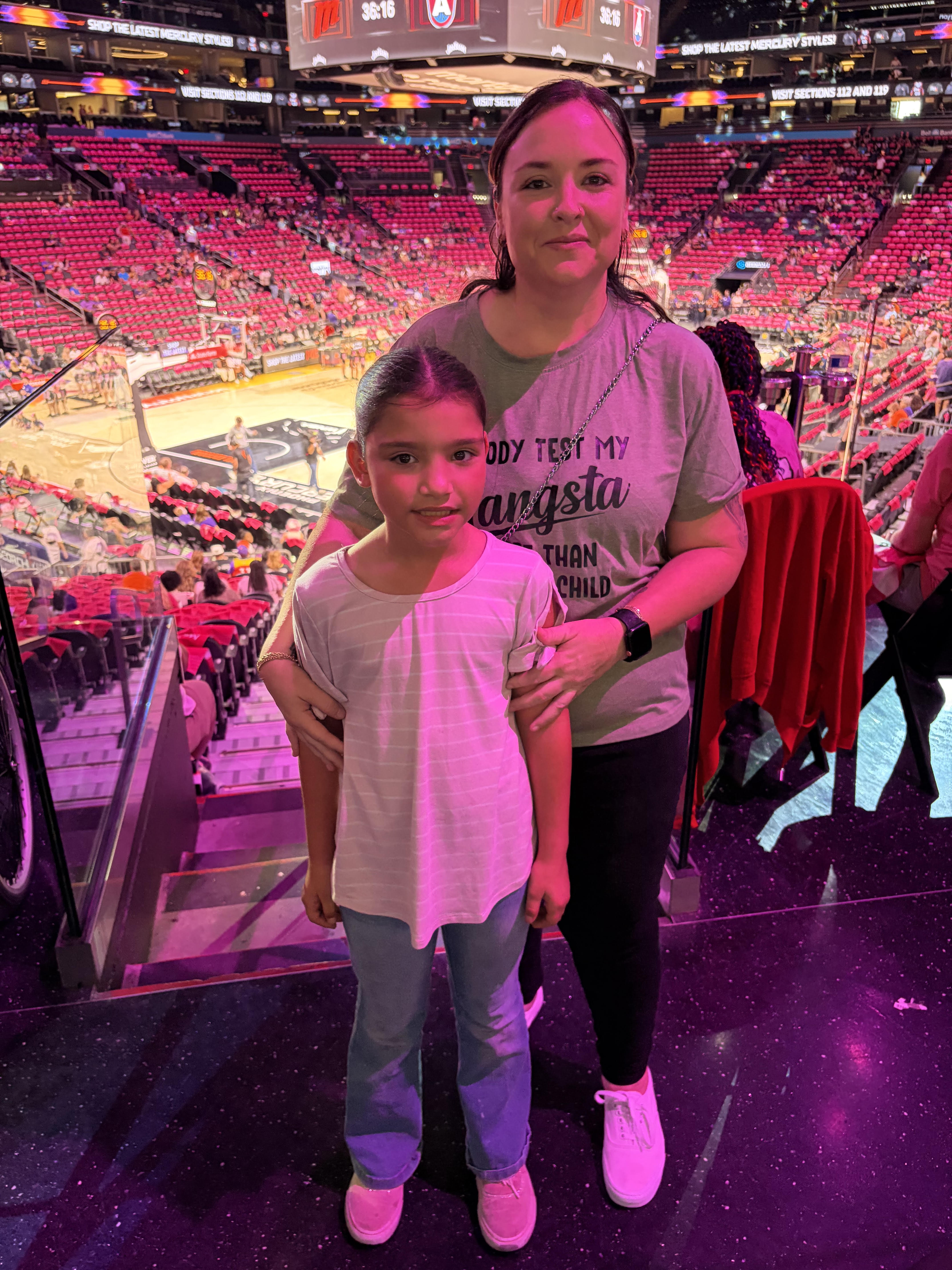 A woman and a young girl stand indoors in an arena with red seats around a basketball court in the background, illuminated by soft pink lighting.