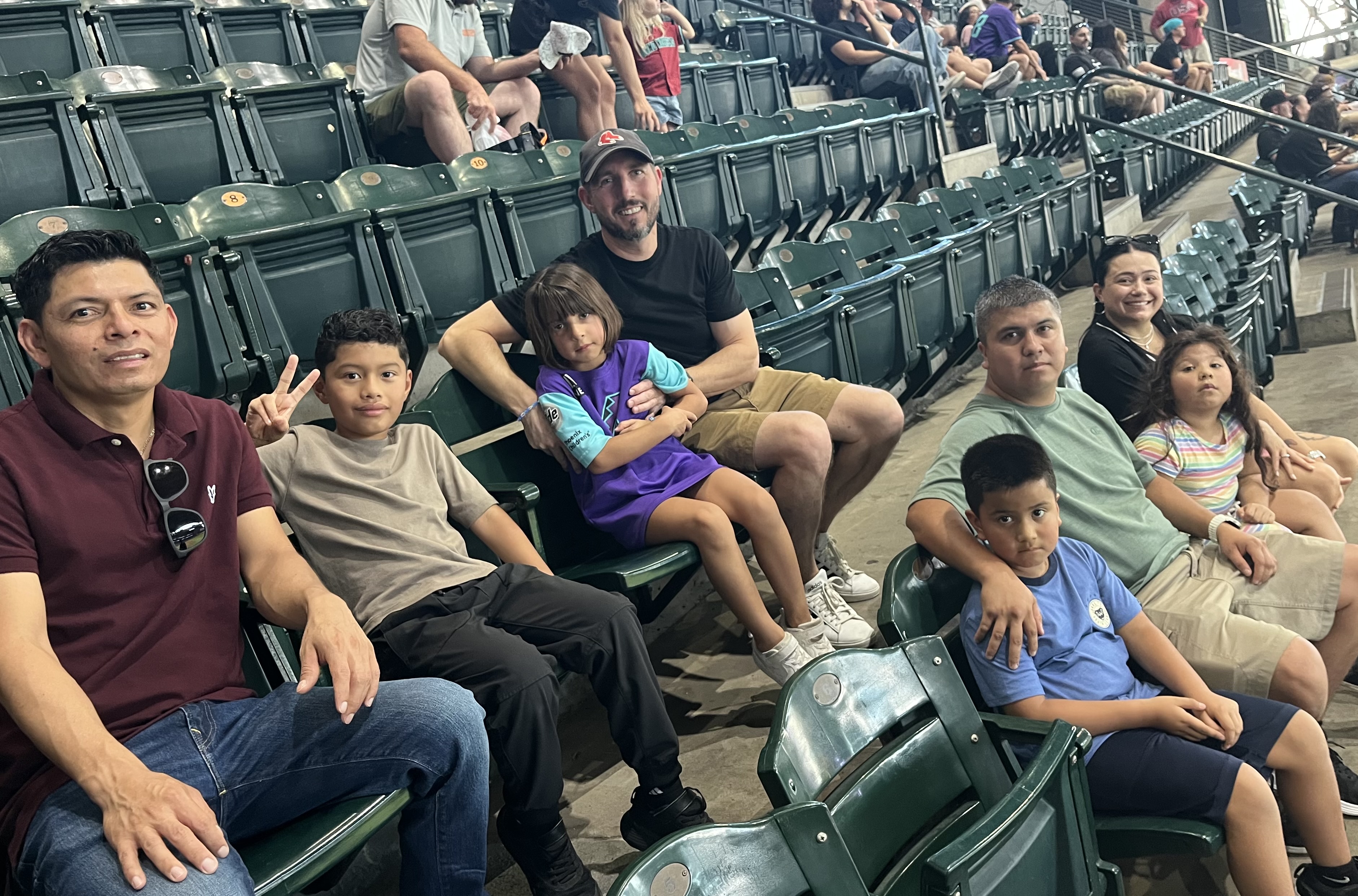 A group of seven people, including four adults and three children, sitting together in green stadium seats at a sports venue, with some smiling and one boy making a peace sign.