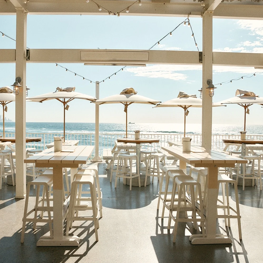 Covered terrace of white high tables and stools, with a row of umbrellas along the railing and the ocean beyond.