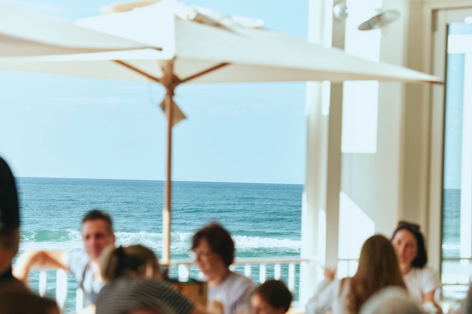 Diners in soft focus beneath a white umbrella, with blue sky and waves rolling in on the ocean beyond.