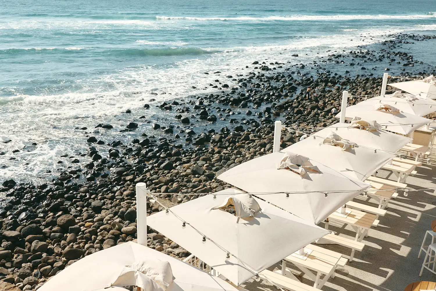 Overhead view of white umbrellas and festoon lights lining the terrace, beside dark boulders and waves washing ashore.