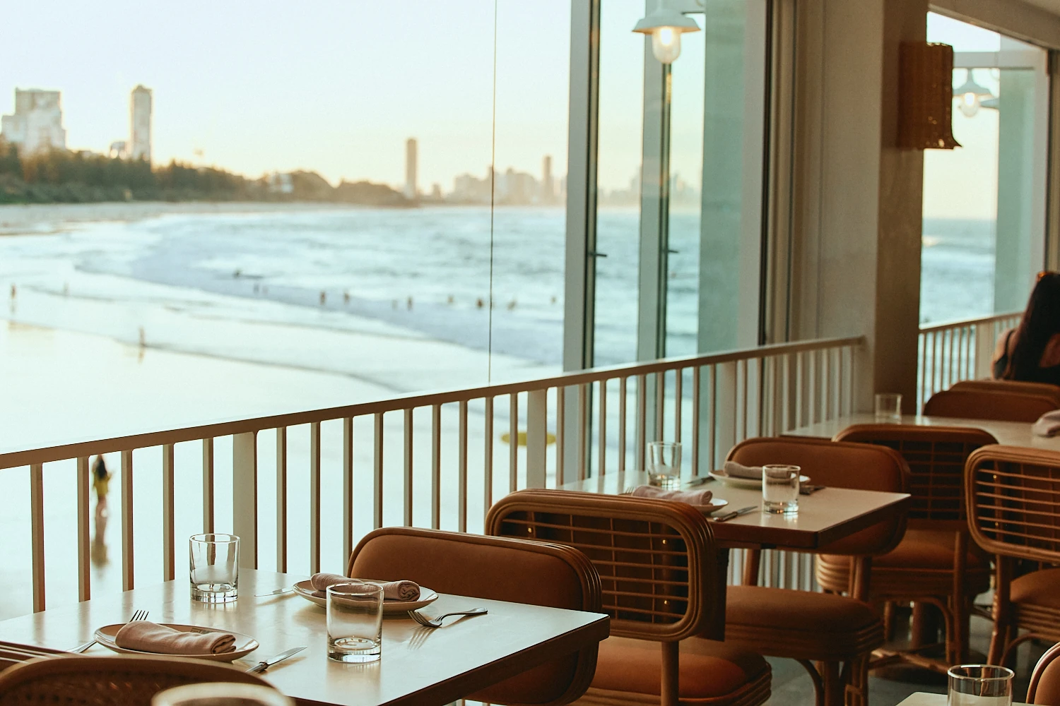 Tables with rattan chairs by the balcony at golden hour, overlooking swimmers on Burleigh Beach and distant high-rises.