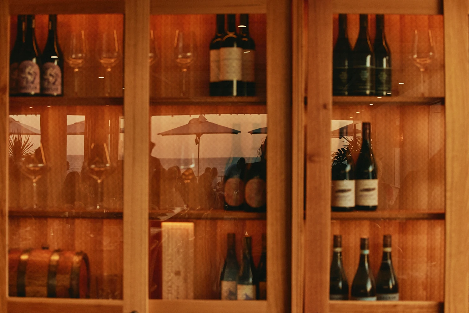 Wine bottles and glasses in a warmly lit timber cabinet, its glass doors reflecting umbrellas and diners on the terrace.