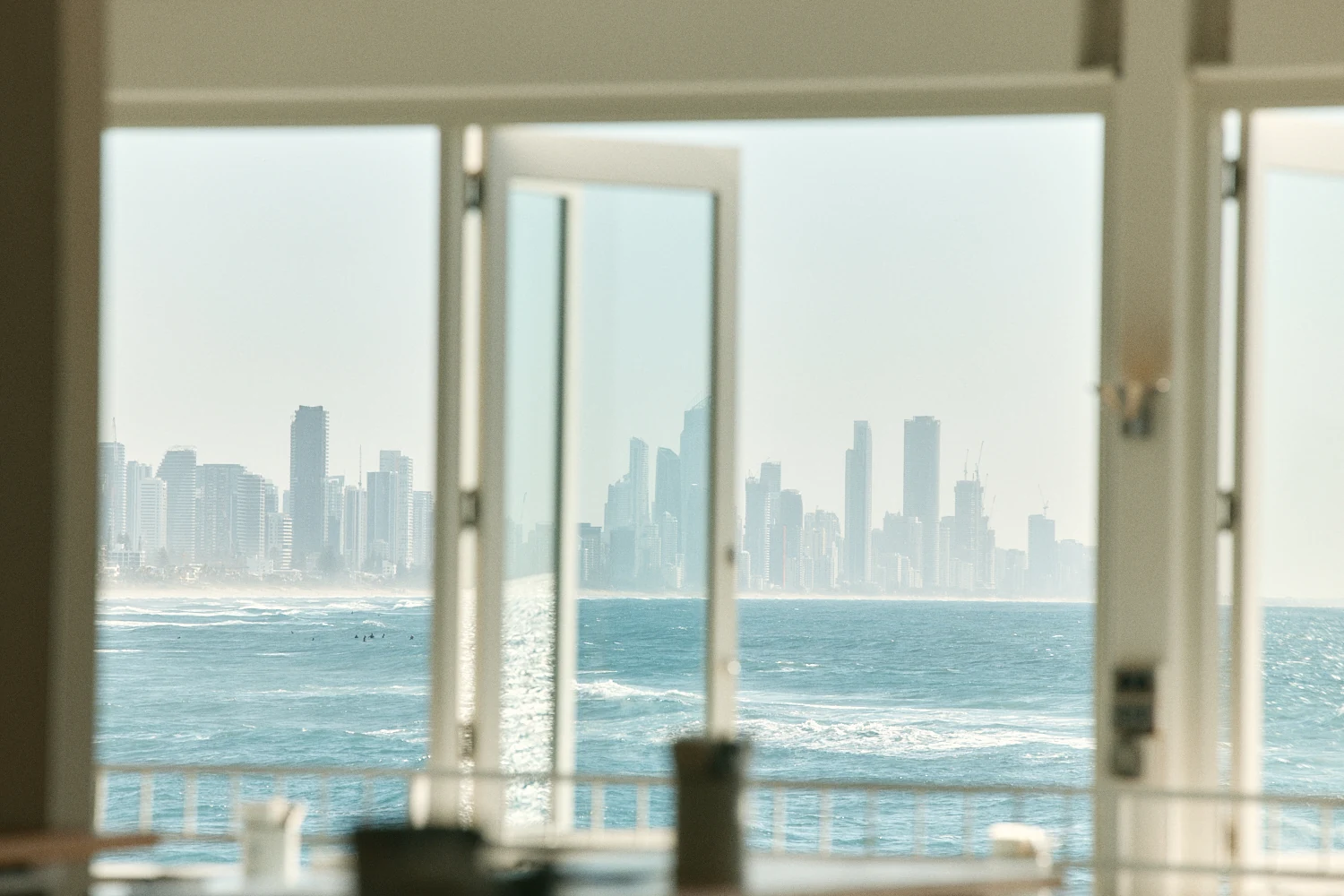 Open white windows framing surfers in the ocean and the hazy Gold Coast skyline across the water.