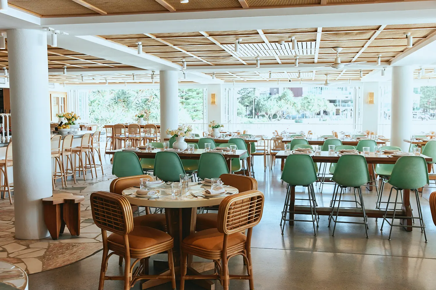 Bright dining room under a woven timber ceiling, with rattan chairs at a round table and mint green stools at high tables.