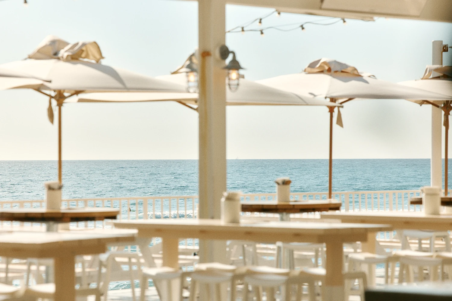 Cream umbrellas over white tables and stools in soft focus, with a calm ocean stretching to the horizon.
