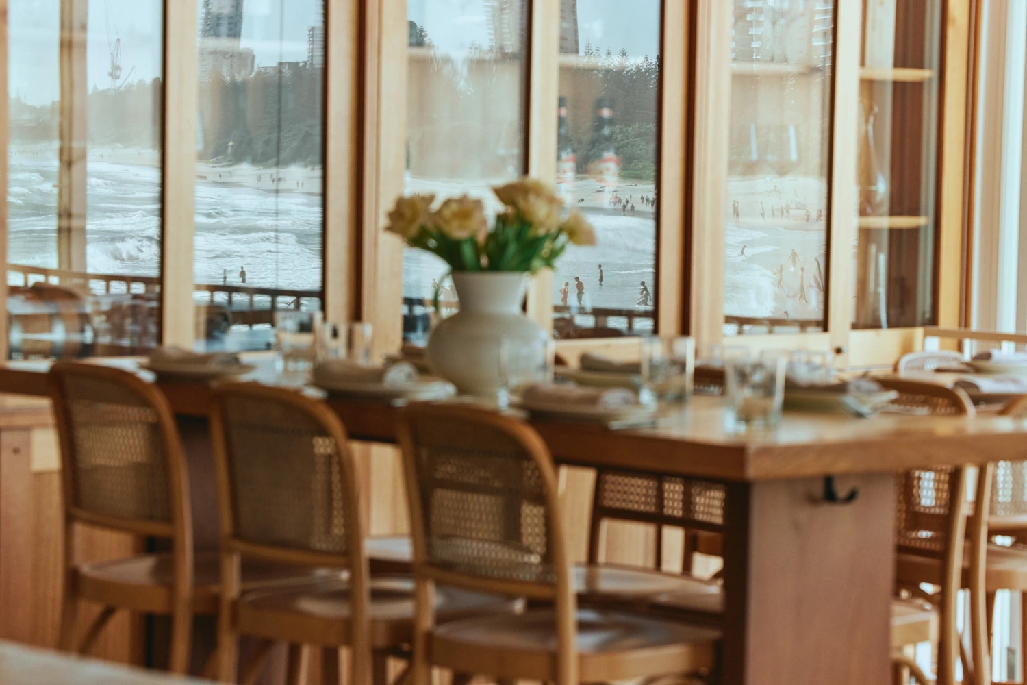Long timber table with cane-back chairs and a vase of pale yellow flowers, in front of windows looking out to the beach.