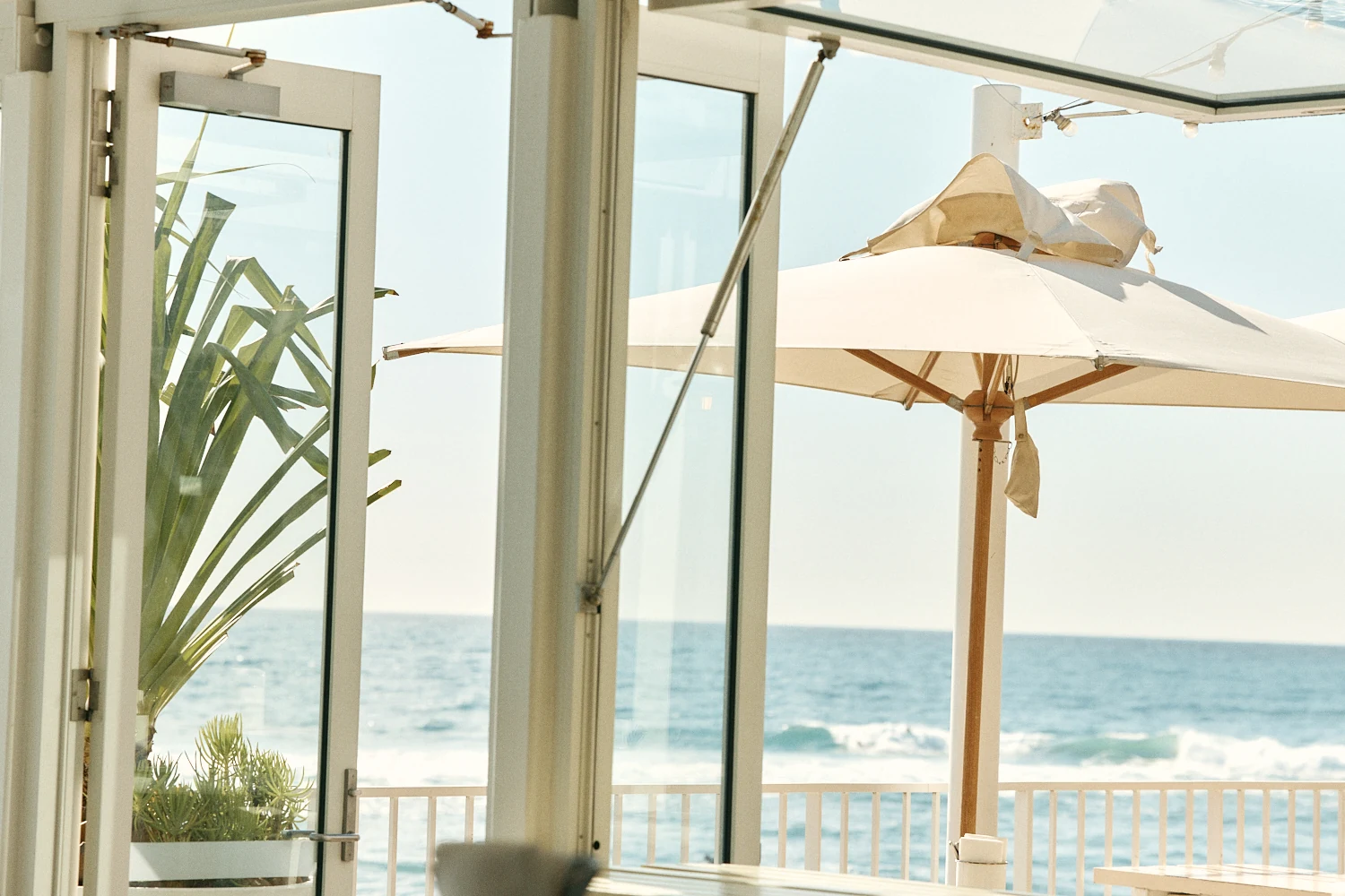 Open glass doors and windows looking out to a white umbrella, a potted plant and waves breaking beyond the railing.