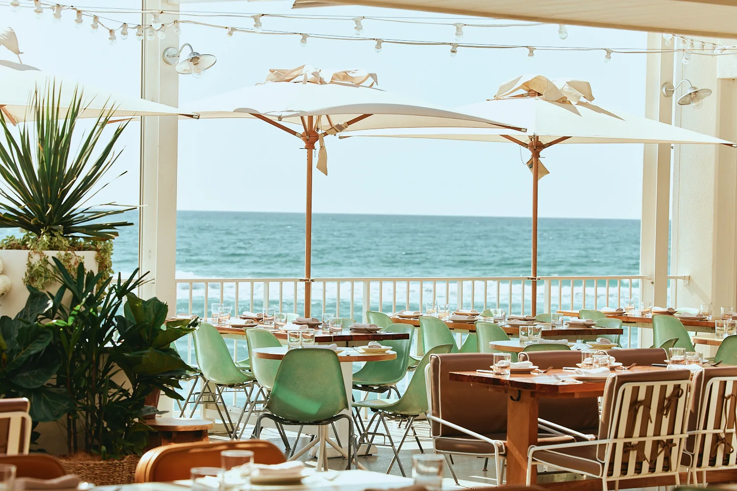 Oceanfront terrace set for dining under white umbrellas and festoon lights, framed by tropical plants.