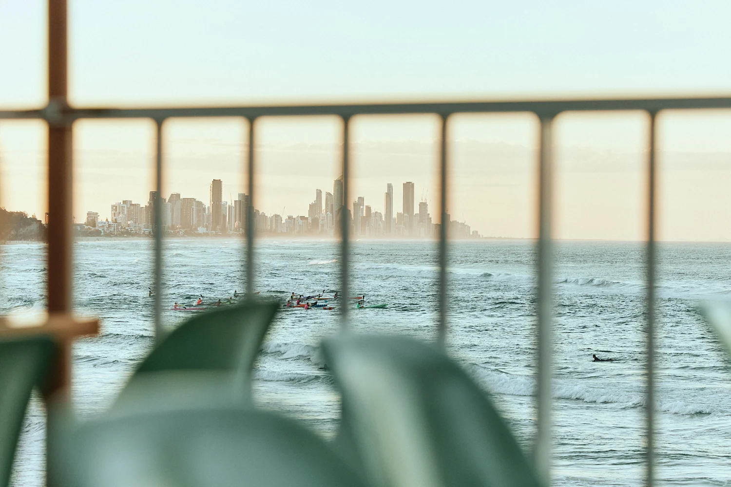 Paddlers and a lone surfer out on the ocean, seen through the balcony railing, with the hazy Gold Coast skyline beyond.