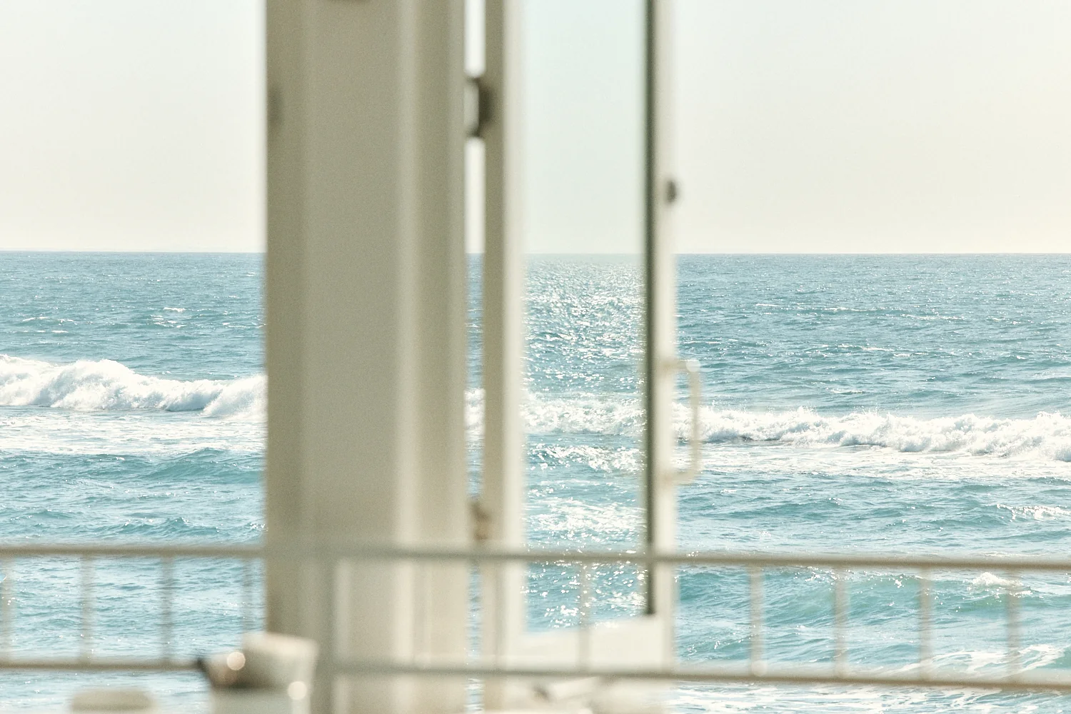 Waves breaking on a sparkling blue ocean, seen past an open white window and railing.