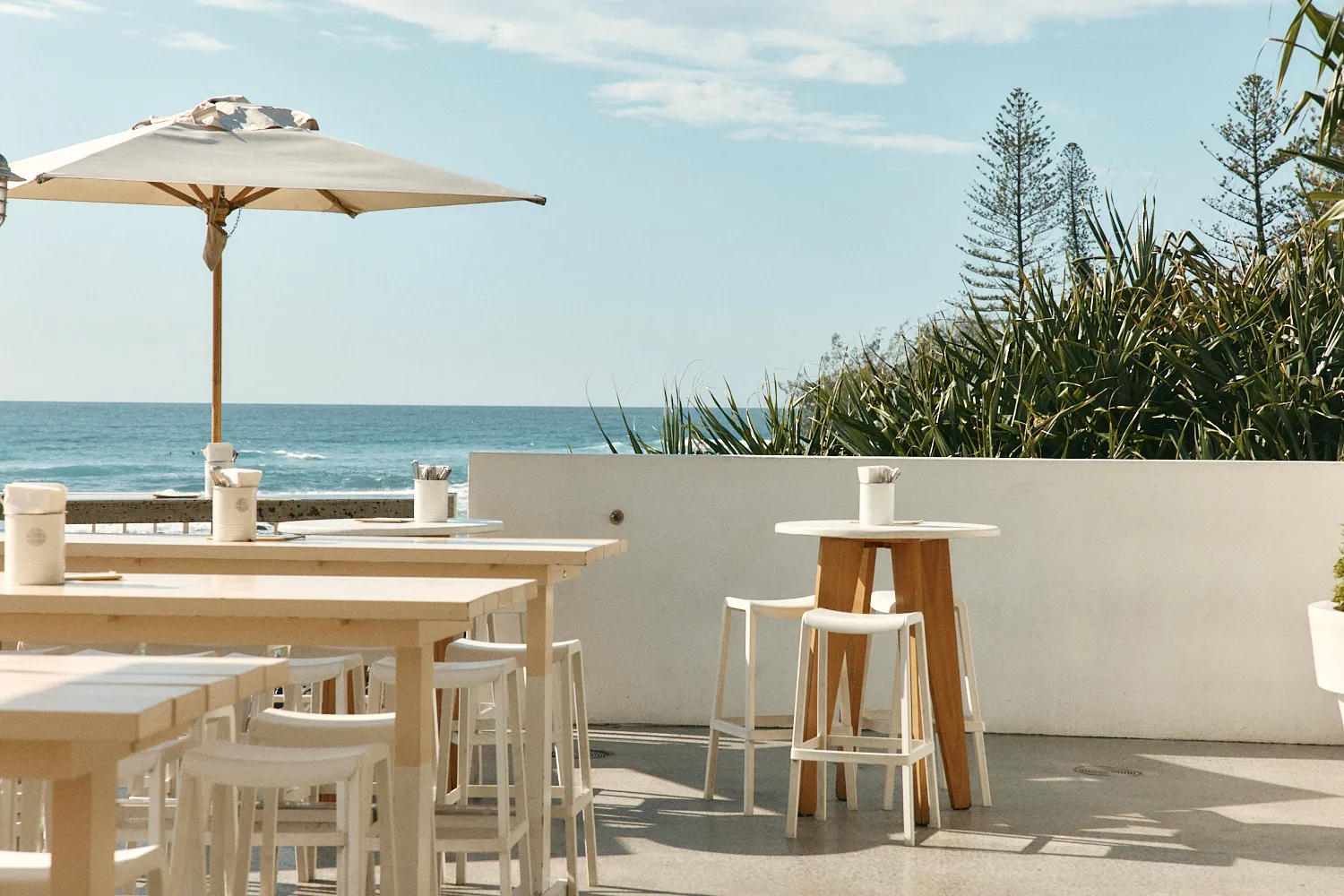White stools at high tables beside a low white wall, with the ocean on one side and pandanus and pines on the other.