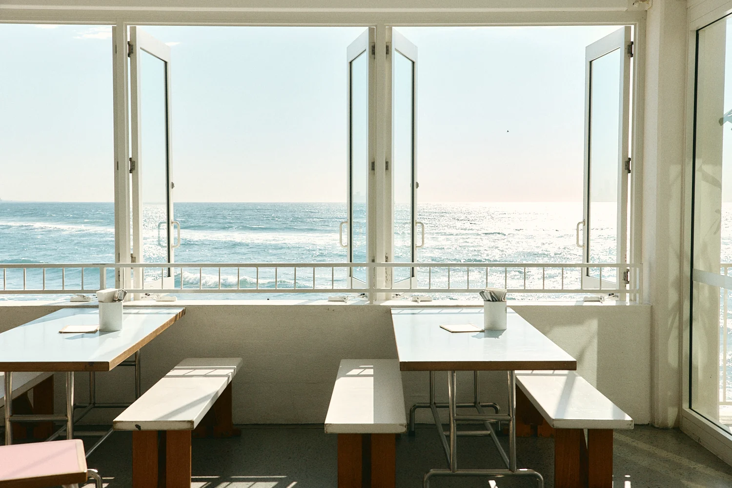 White tables and timber benches beneath open windows, looking out over the railing to a sunlit ocean.
