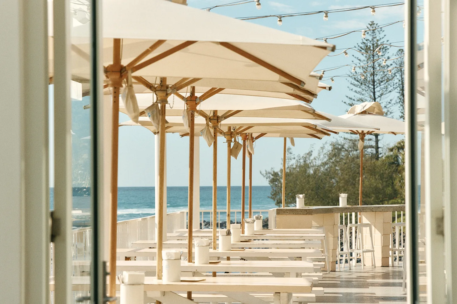 Through open doors, a long row of white tables under timber-pole umbrellas and festoon lights, with the ocean beyond.