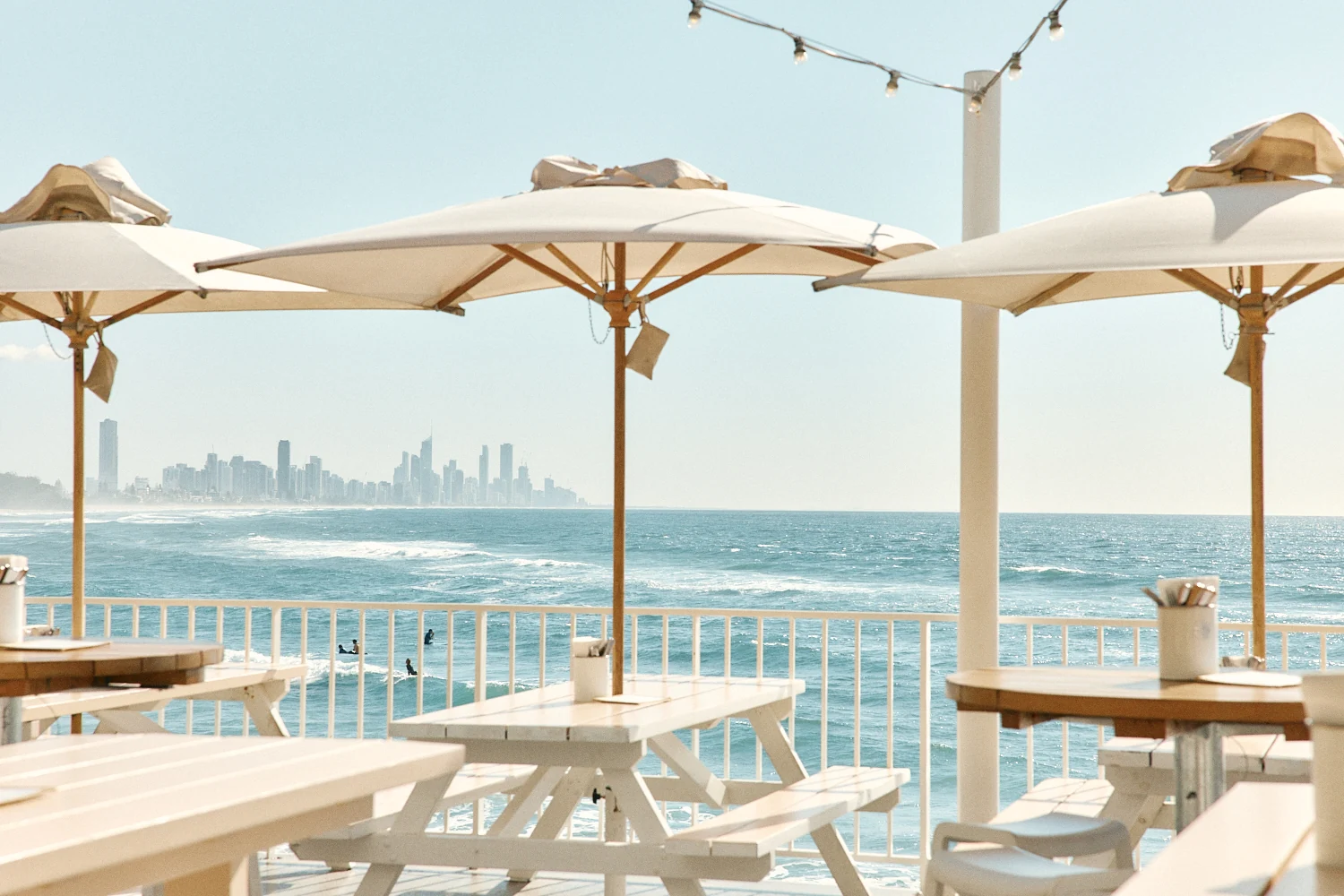 White picnic tables under umbrellas by the railing, with surfers in the waves and the Gold Coast skyline beyond.