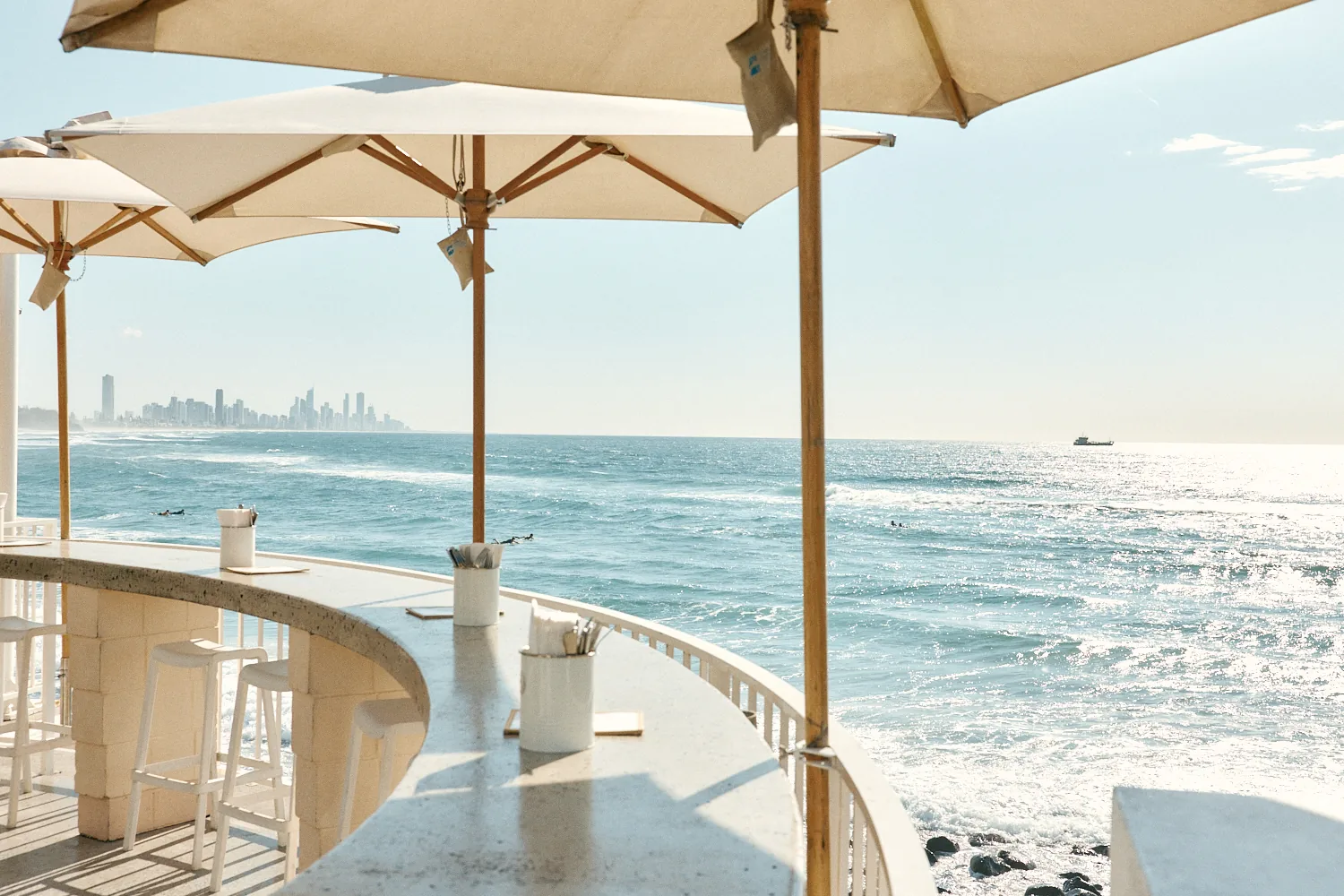 Curved concrete bar ledge with stools along the railing, overlooking the sparkling ocean, surfers and a distant skyline.