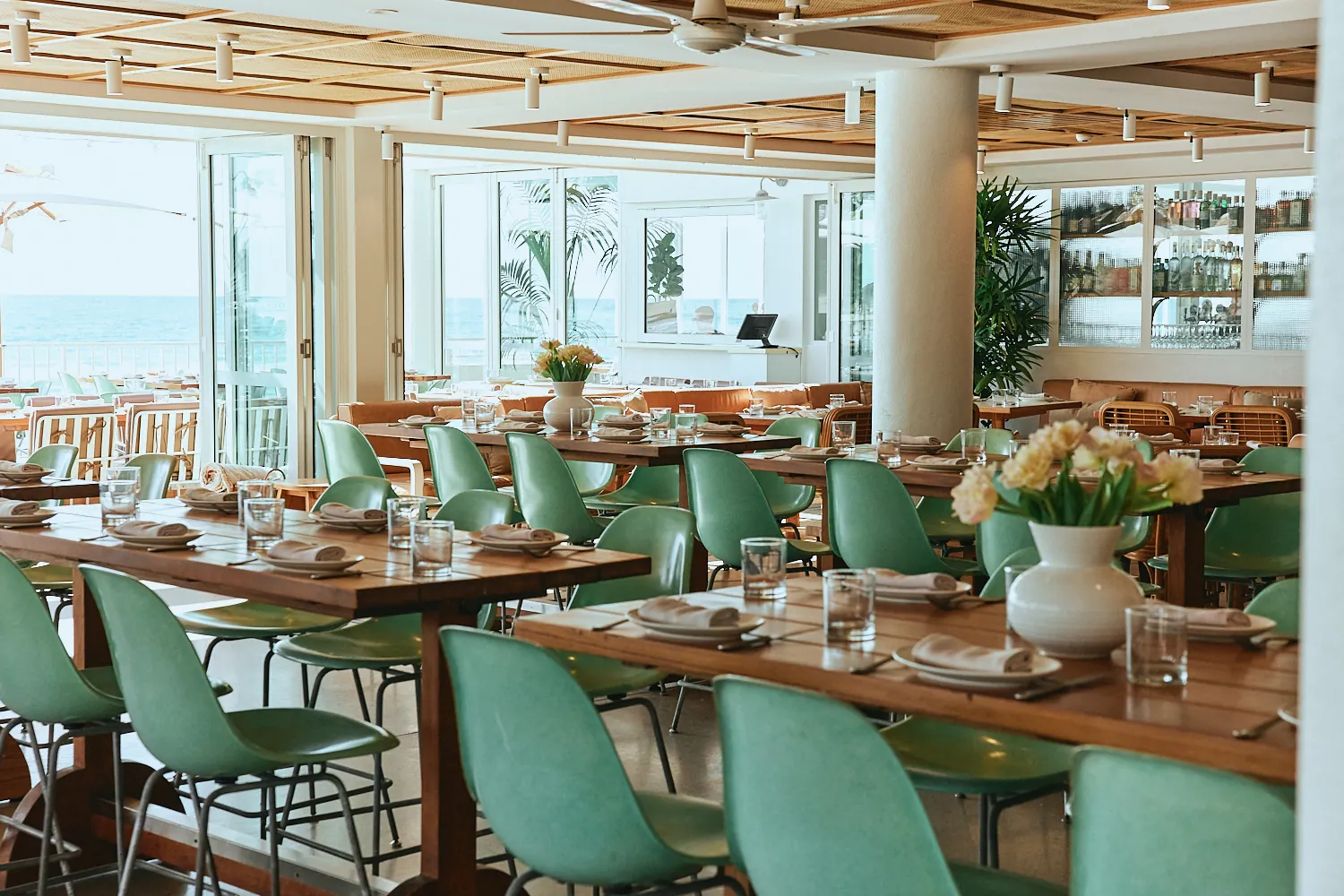 Mint green chairs at long timber tables, with doors open to the ocean terrace and a back bar lined with bottles.