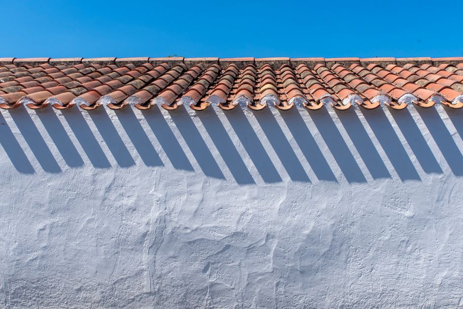 A terracotta tiled roof casting shadows on a white wall under a clear blue sky.