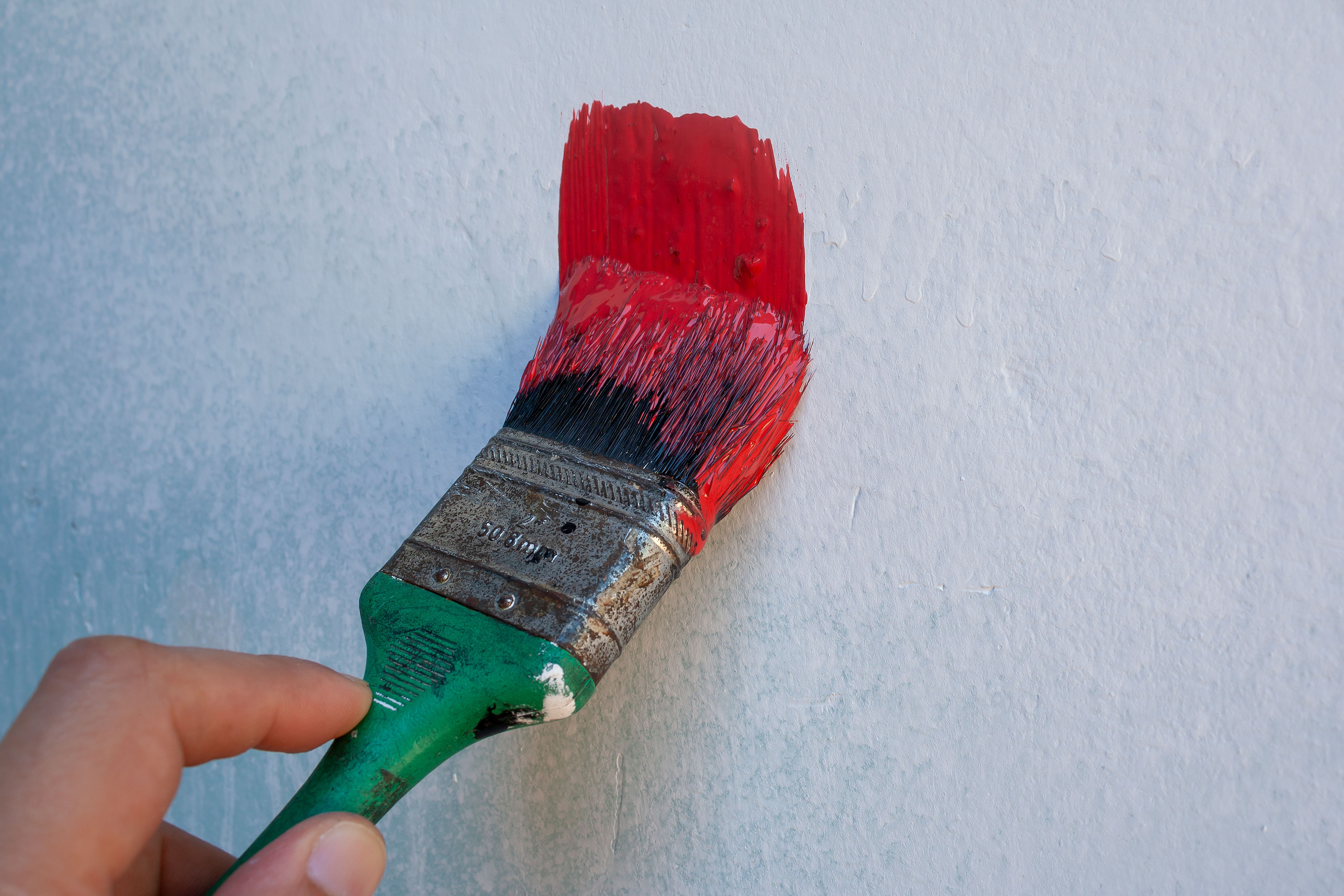 Detailed image of a hand holding a paintbrush with red paint on a textured wall.