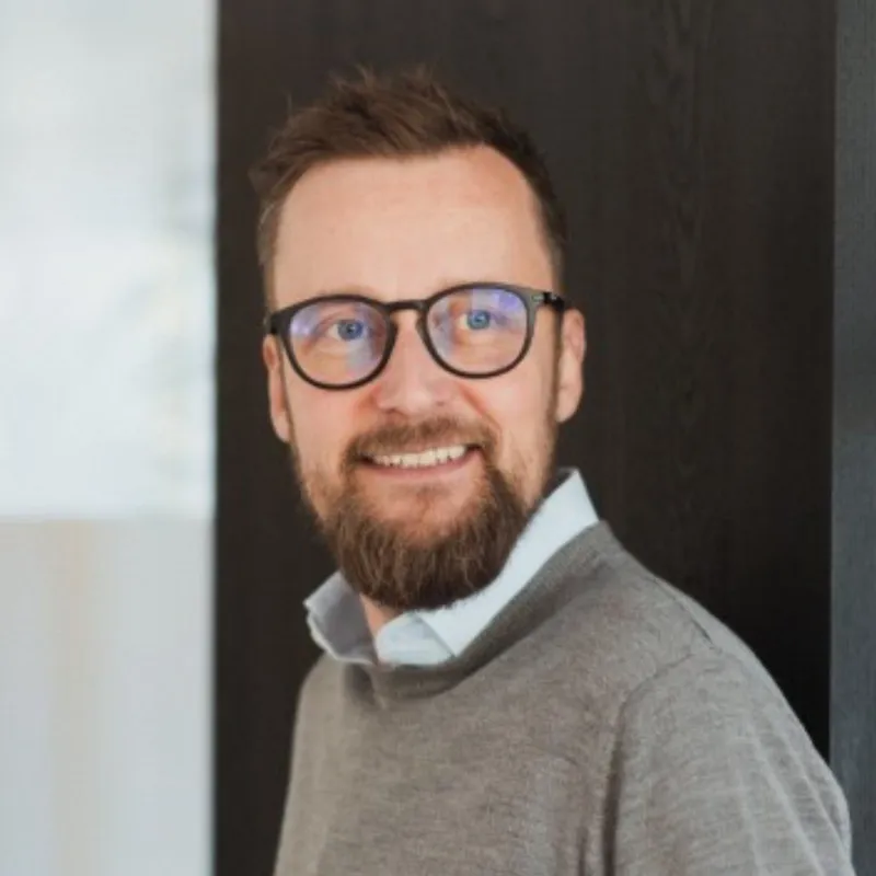 Smiling man with glasses, a beard, and short hair wearing a gray sweater over a light blue collared shirt standing indoors against a dark background.