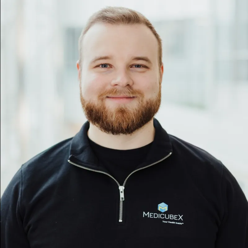 Smiling bearded man with light hair wearing a black zip-up top with the MedicubeX logo and slogan 'Your Health Inside'.