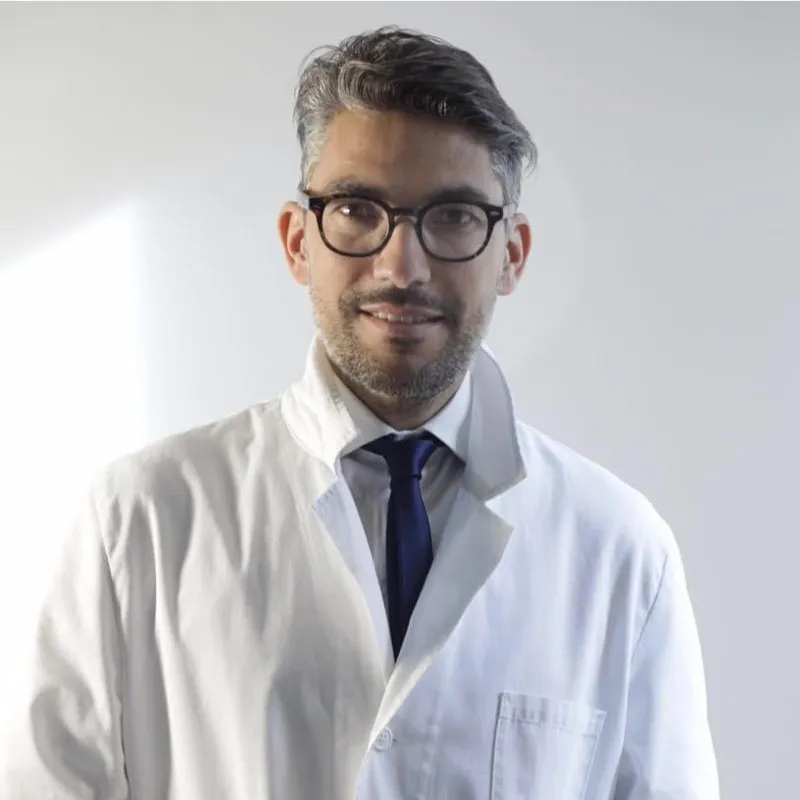Man with dark hair and glasses wearing a white lab coat and dark tie, smiling slightly against a plain light background.