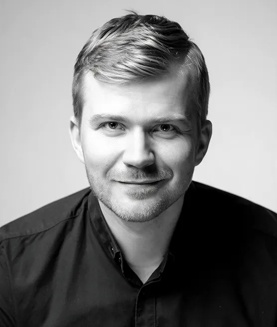 Black and white portrait of a young man with short, styled hair and light stubble wearing a dark collared shirt, smiling slightly against a plain background.