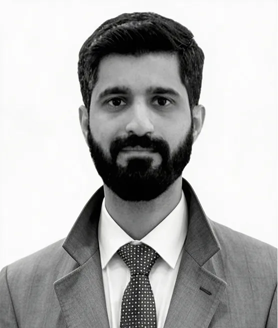 Portrait of a man with dark hair and beard wearing a suit, white shirt, and patterned tie against a plain background.