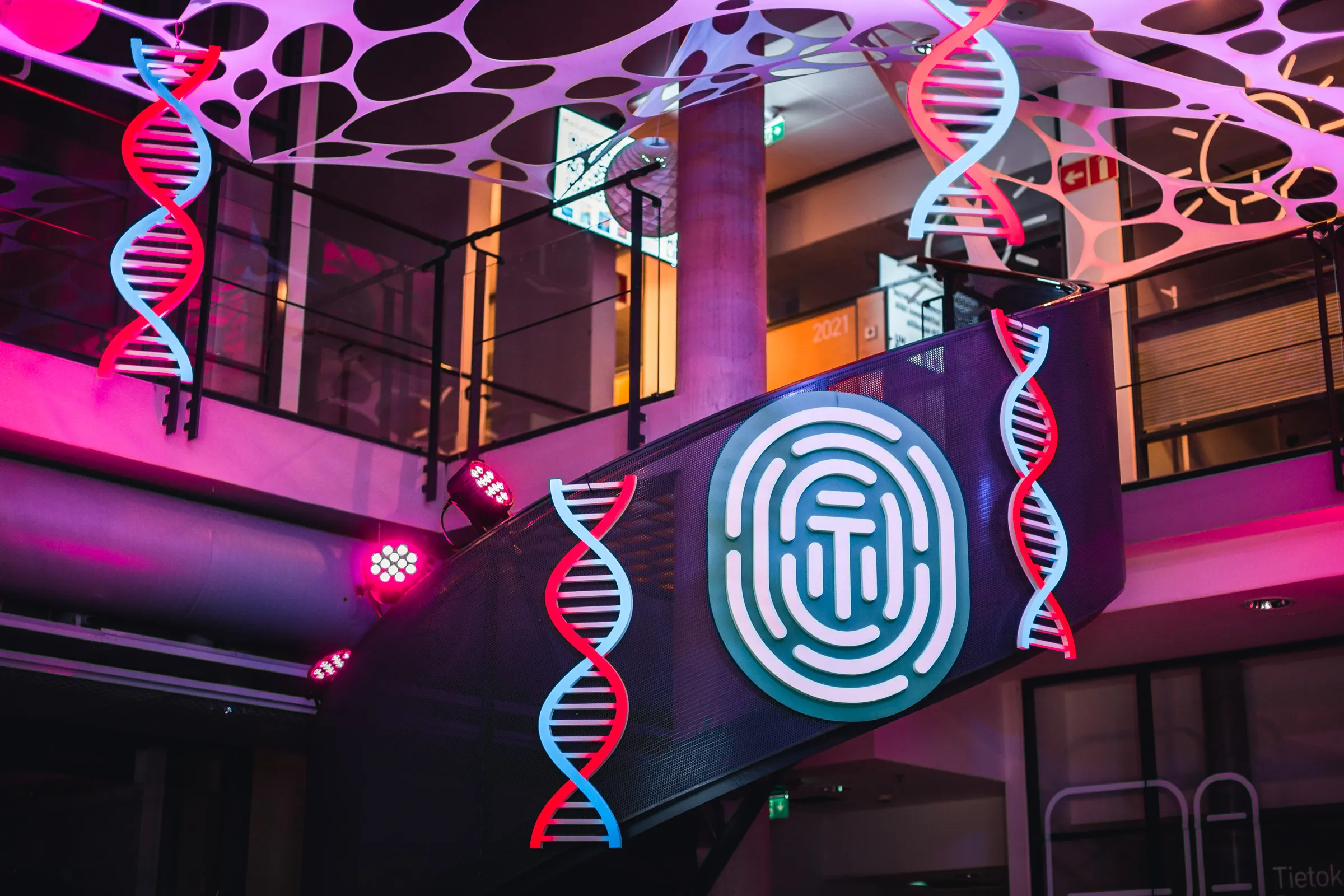 Modern interior featuring large illuminated DNA double helix models hanging from the ceiling and a glowing fingerprint pattern on a black mesh wall, with purple and pink accent lighting.
