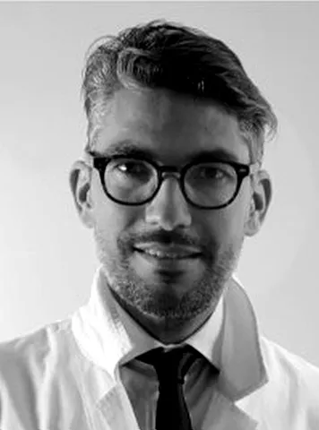 Man with short, neatly styled hair and beard wearing round glasses, a white lab coat, white shirt, and black tie, against a plain light background.