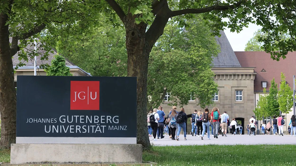 Outdoor scene at Johannes Gutenberg University Mainz showing a large sign with the university name and logo in front of green trees, with students walking on a pathway towards a historic building with stone architecture and red-tiled roof in the background.