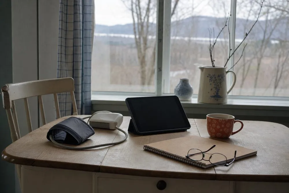 Wooden table near window with a blood pressure monitor, a tablet on stand, a patterned orange coffee mug, a notebook, and reading glasses arranged on it, with a chair on the left and a jug with branches and a small vase on the windowsill in the background.
