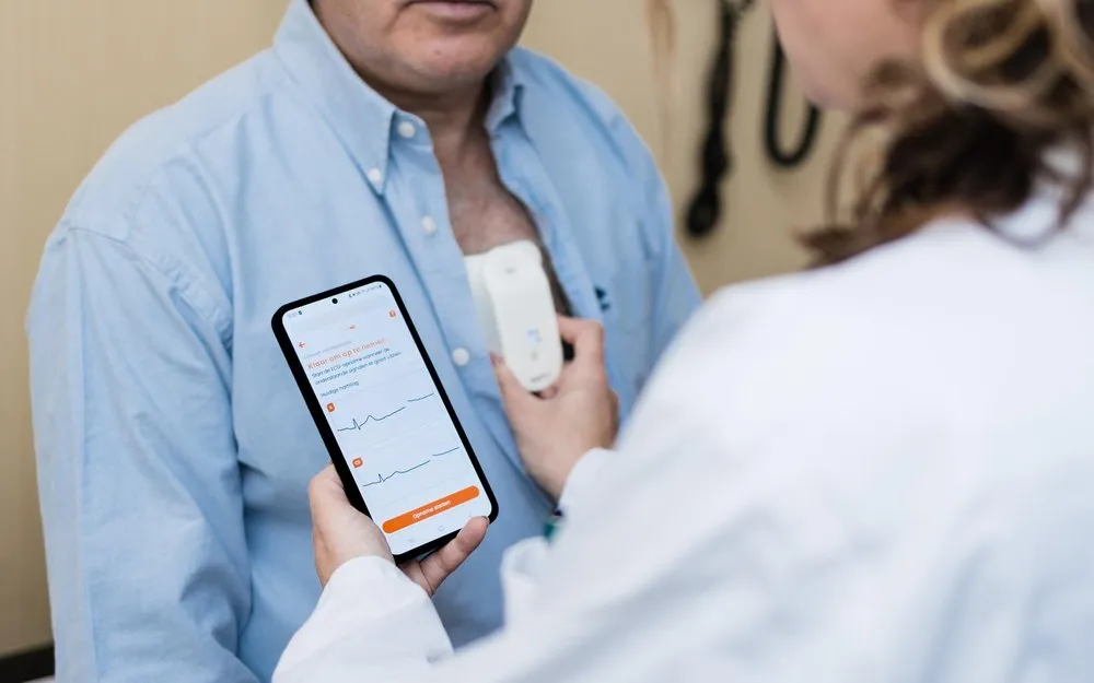 A healthcare professional holding a smartphone showing ECG readings while attaching a white ECG patch device to a man's chest, who is wearing a blue shirt.