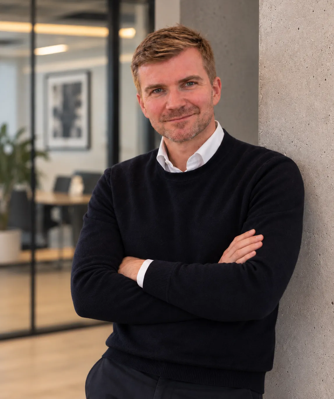 Smiling man with short blonde hair and light beard wearing a white collared shirt under a black sweater, standing with arms crossed leaning against a concrete wall in a modern office space.