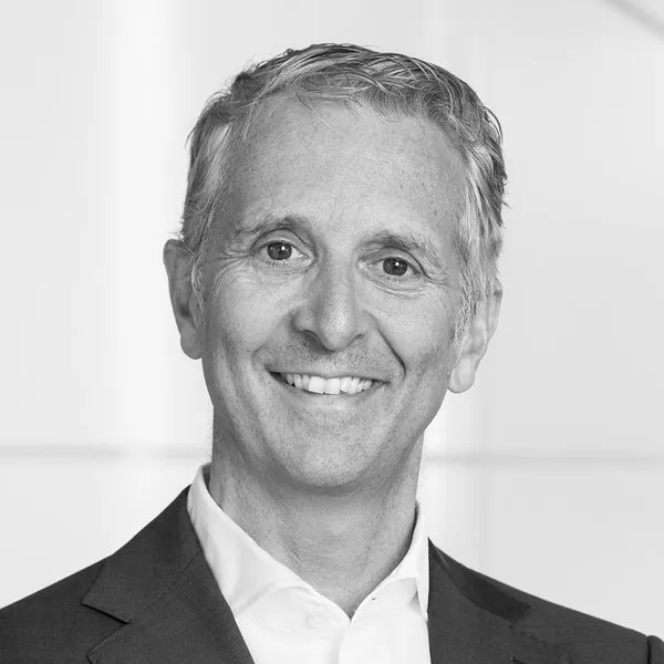 Smiling middle-aged man with short gray hair wearing a dark suit and white shirt, against a light blurred background.