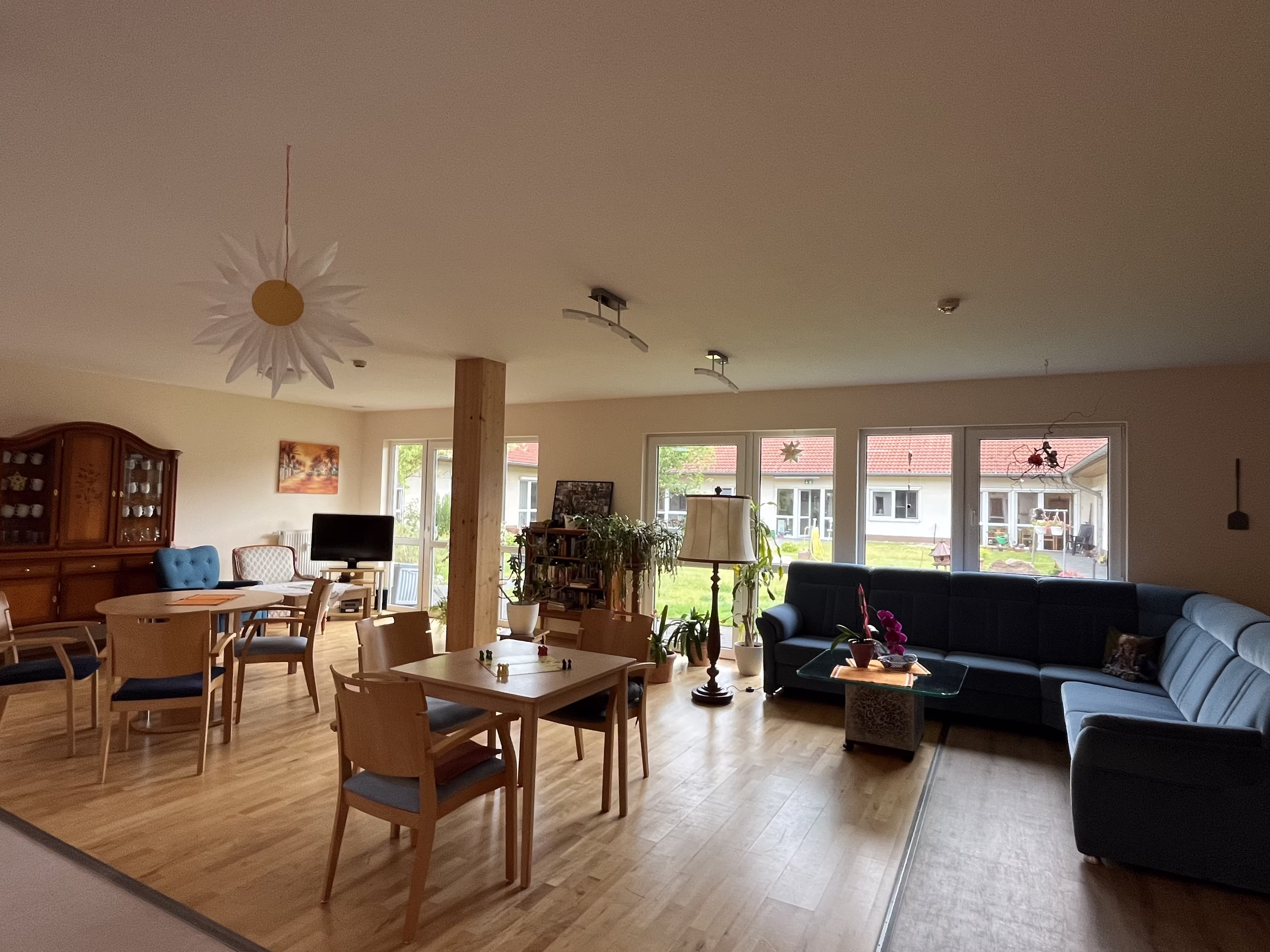 Helle Gemeinschaftsfl&auml;che des Seniorenzentrums mit langer Tafel, gem&uuml;tlicher Sofaecke und gro&szlig;er Fensterfront zum Innenhof
