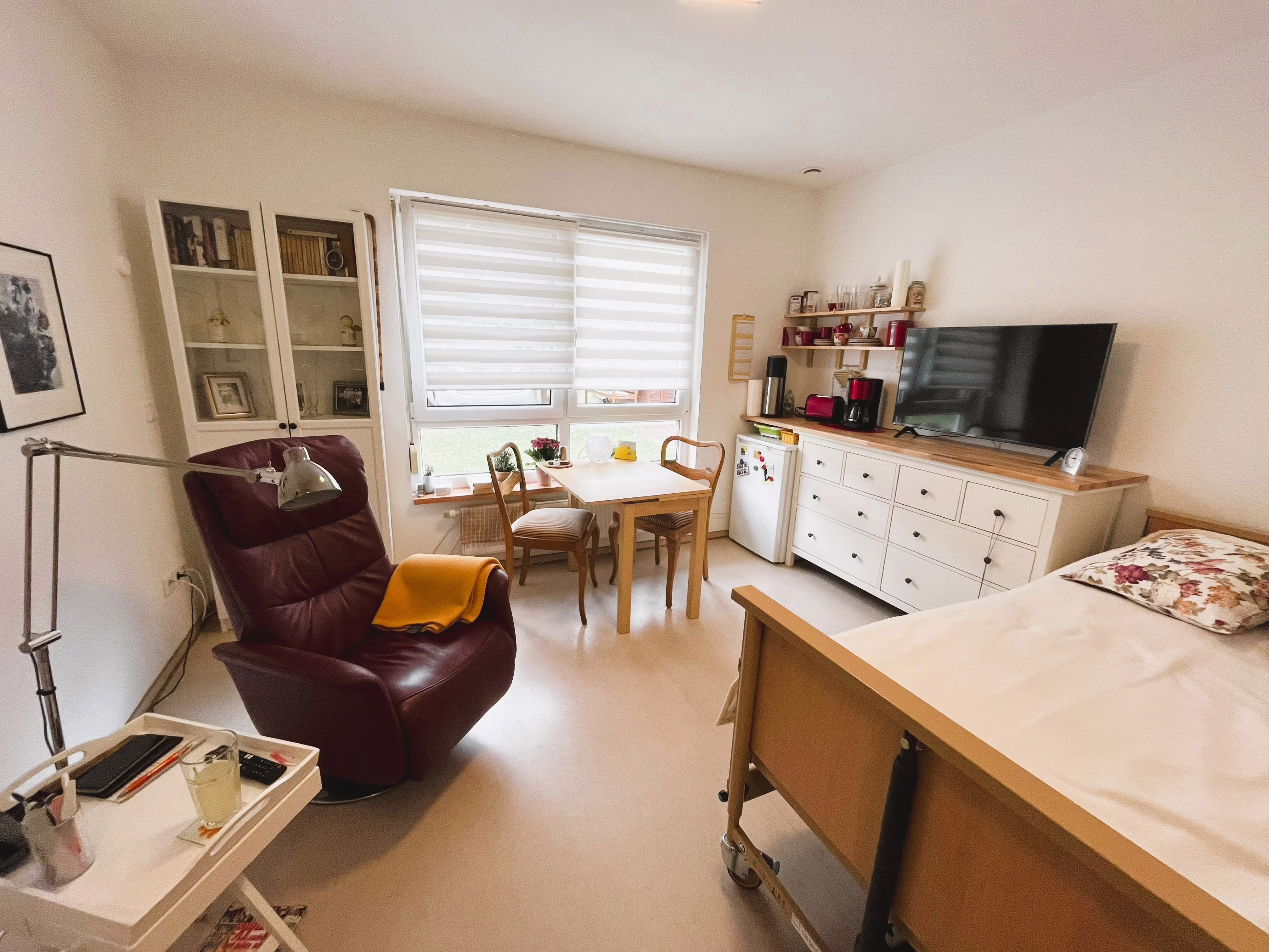 Apartment im Betreuten Wohnen Plus mit bequemem Sessel, Essplatz am Fenster, Kommode und h&ouml;henverstellbarem Pflegebett