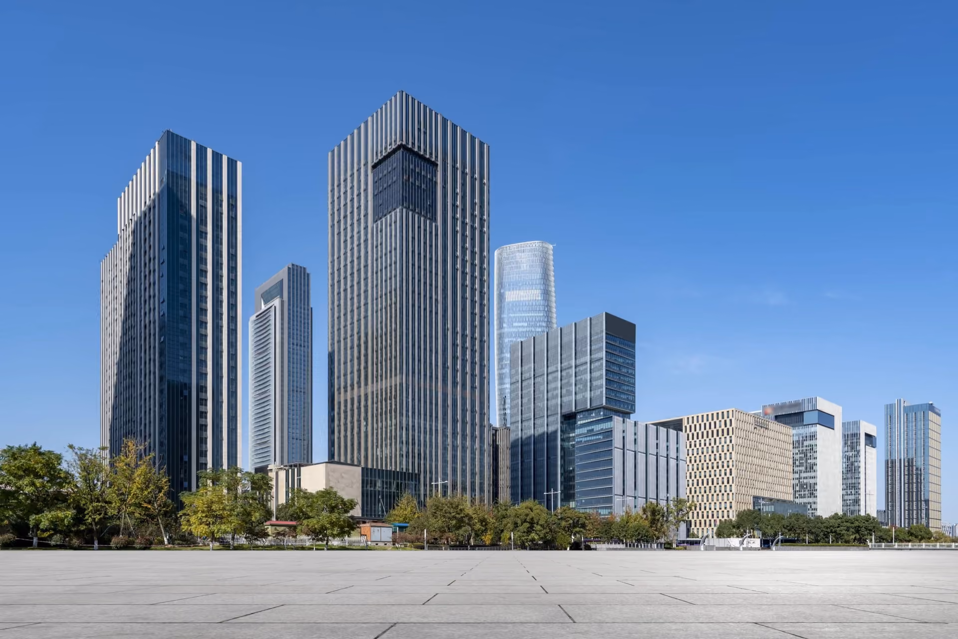 Dark glass high-rises and a line of trees beyond an open stone plaza