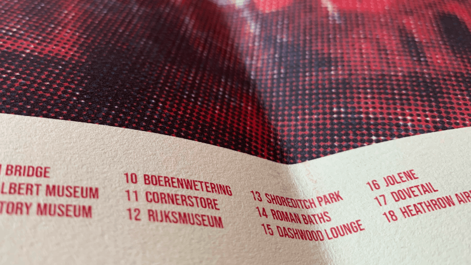 Close-up photo of printed text in red on textured beige paper listing numbered locations including Boerenwetering, Cornerstore, Rijksmuseum, Shoreditch Park, Roman Baths, Dashwood Lounge, Jolene, Dovetail, and Heathrow Airport.