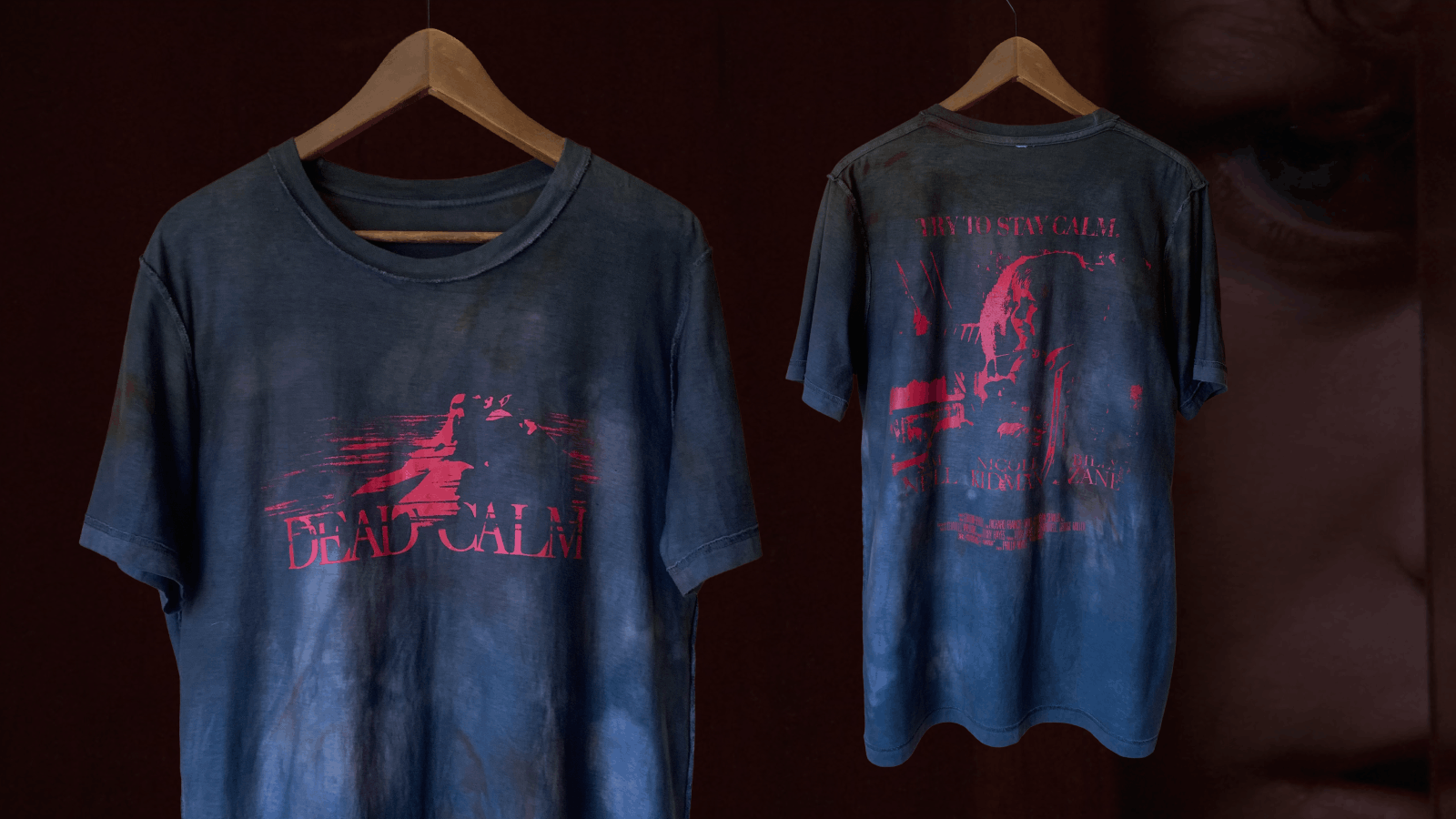 Two faded navy blue t-shirts hanging on wooden hangers against a dark background; the front shirt features a red graphic with a silhouette and the text 'DEAD CALM', the back shirt has a red graphic with a face and the text 'TRY TO STAY CALM' along with additional smaller text and names.