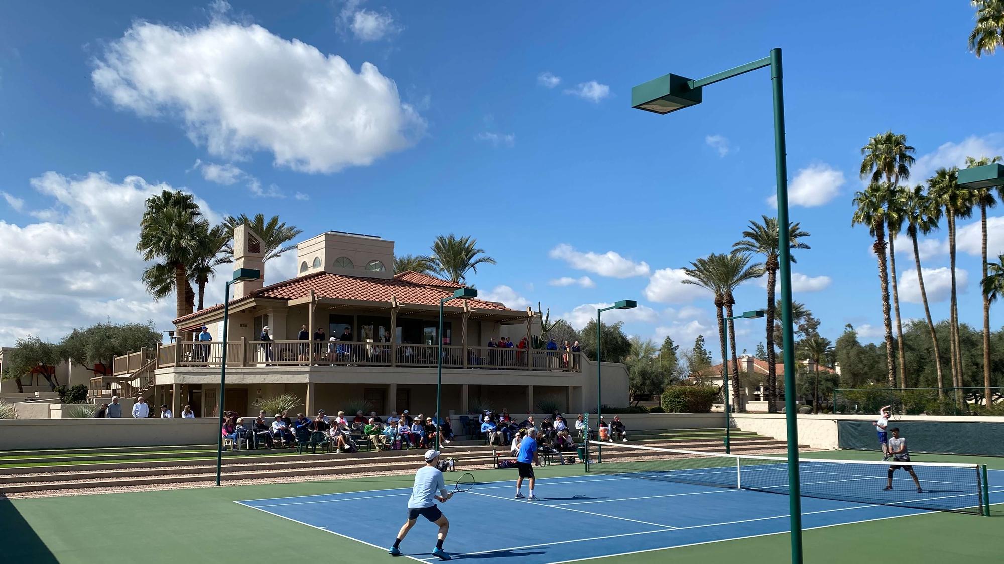 The Racquet Club clubhouse and championship tennis court, with players on court and palm trees beyond.