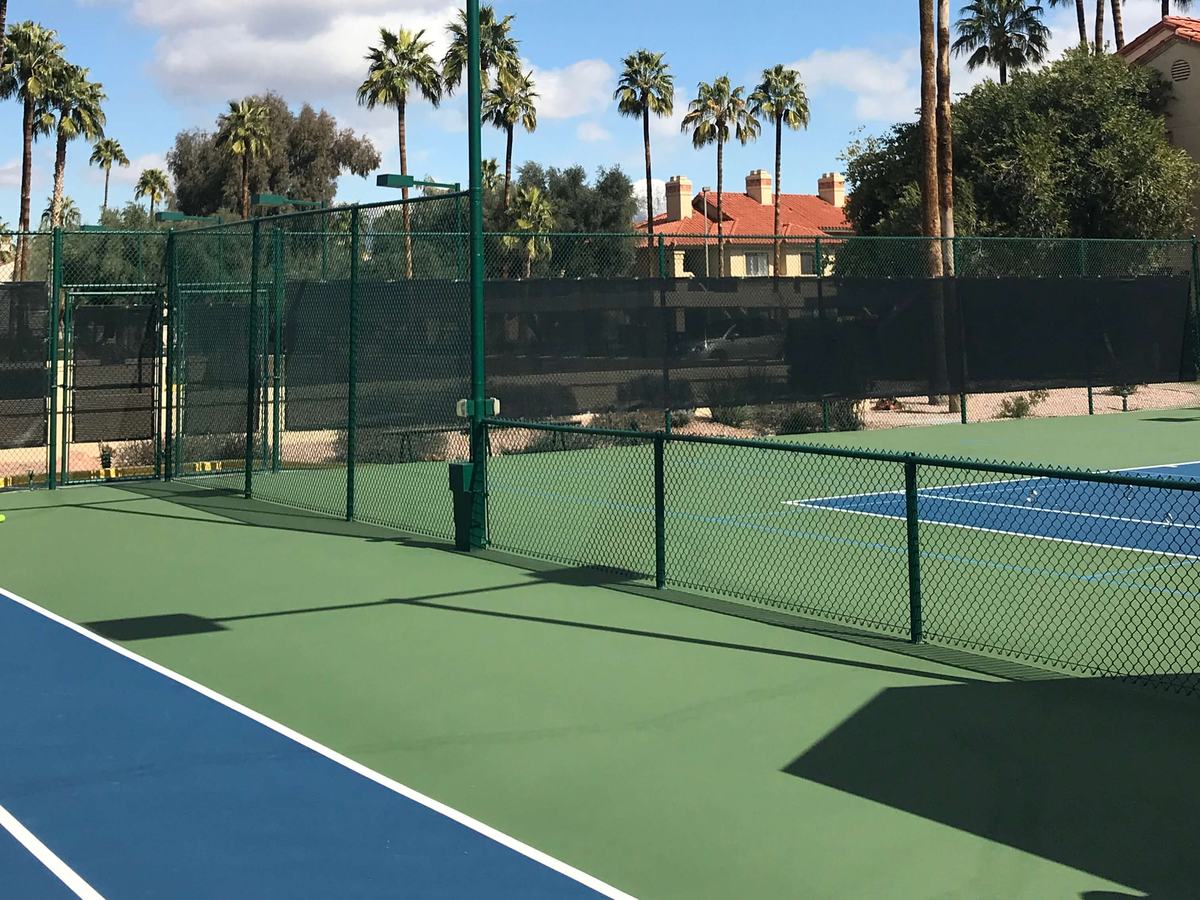 Lighted tennis courts at Racquet Club with palm trees and community rooftops beyond the fence.