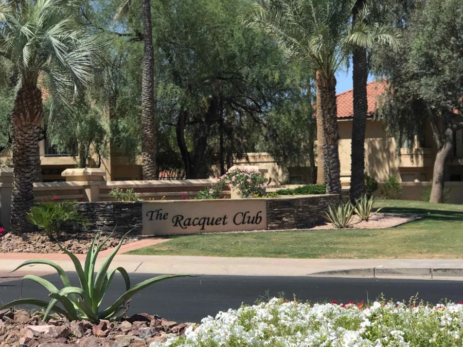 The Racquet Club entrance sign surrounded by flowering plants and palm trees.