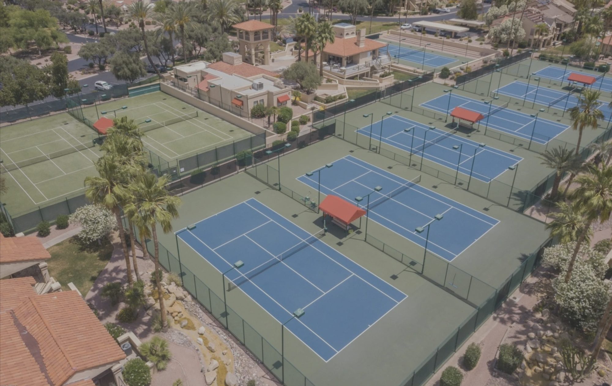 Aerial view of the Racquet Club tennis courts, showing blue hard courts, grass courts, shaded seating shelters and the clubhouse beyond.