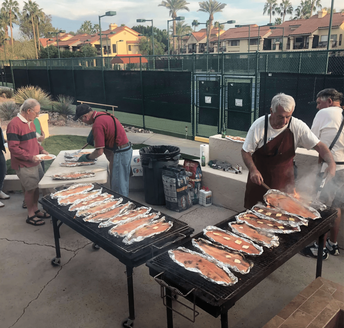 Volunteers preparing trays of food beside an outdoor grill near the tennis courts.