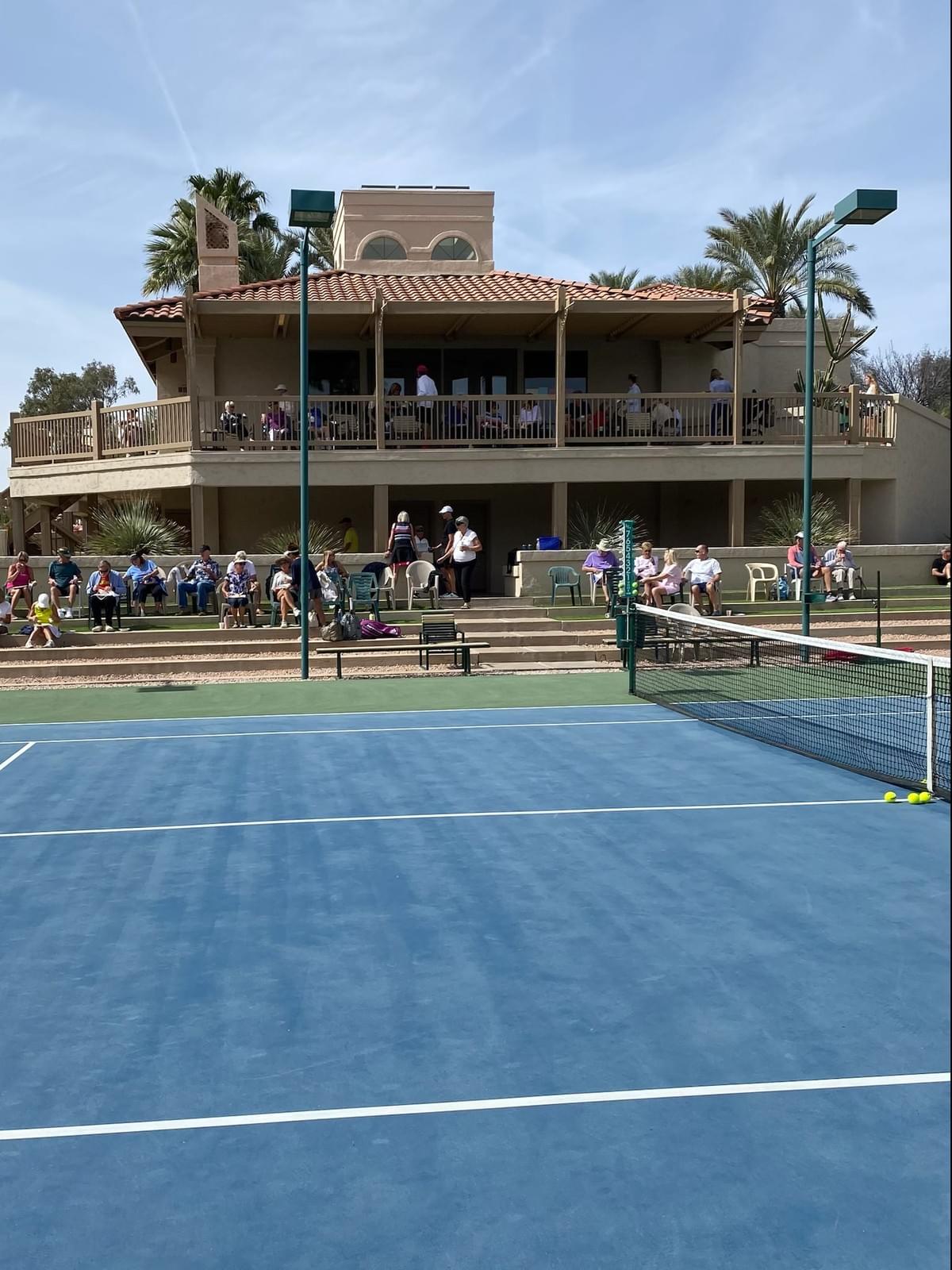 View across the blue tennis court toward spectators and the clubhouse.
